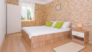 A bedroom with a stone wall, a bed with white bedding and green pillows, a bedside table with a lamp, and a small wooden nightstand.