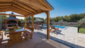 A covered outdoor area with a barbecue, poolside seating, and a stone patio.