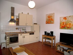 Una cocina compacta con estufa, horno y lavamanos, un comedor con mesa y sillas, y un mueble para televisión con libros.