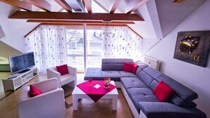 A modern living room with a grey sofa, white armchairs, and a red coffee table. A television is on a white stand, and there is a window with curtains.