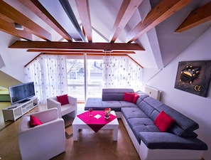 A modern living room with a grey sofa, white armchairs, and a red coffee table. A television is on a white stand, and there is a window with curtains.