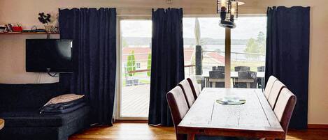 A dining area with a wooden table and chairs, a black sofa, a flat-screen TV, and a view of a building and trees through large windows.