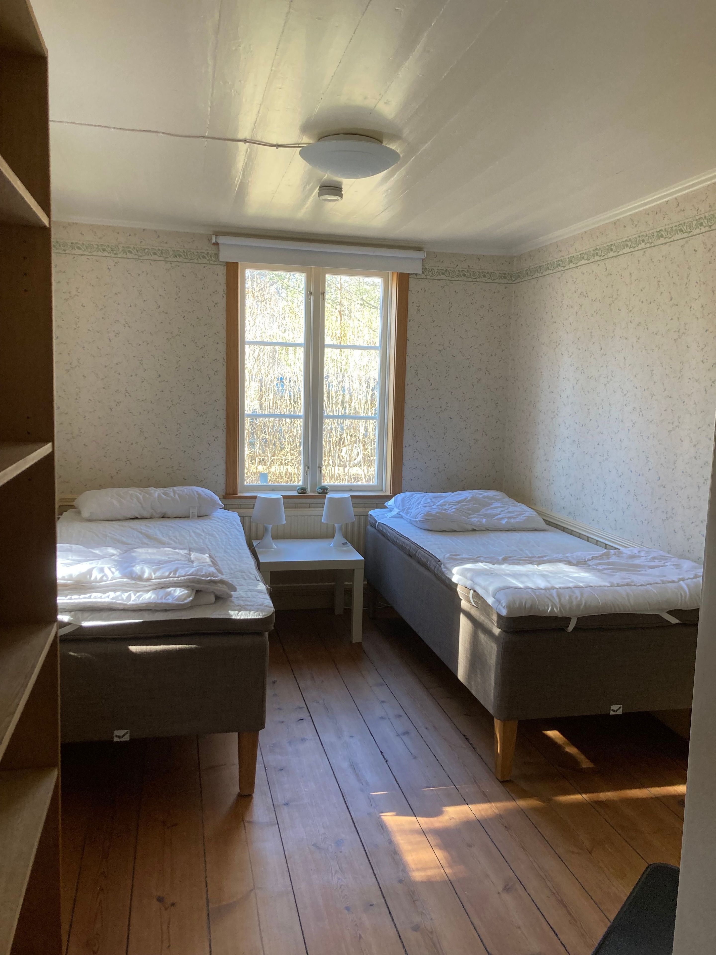 A room with two single beds, a small table, a window, and a ceiling light fixture.