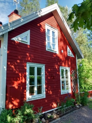 A red wooden house with white trim, a chimney, and a small garden area with plants and a fence.