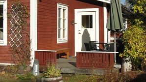 Et rødt træhus med en hvid dør, en lille veranda med en bænk og en parasol.
