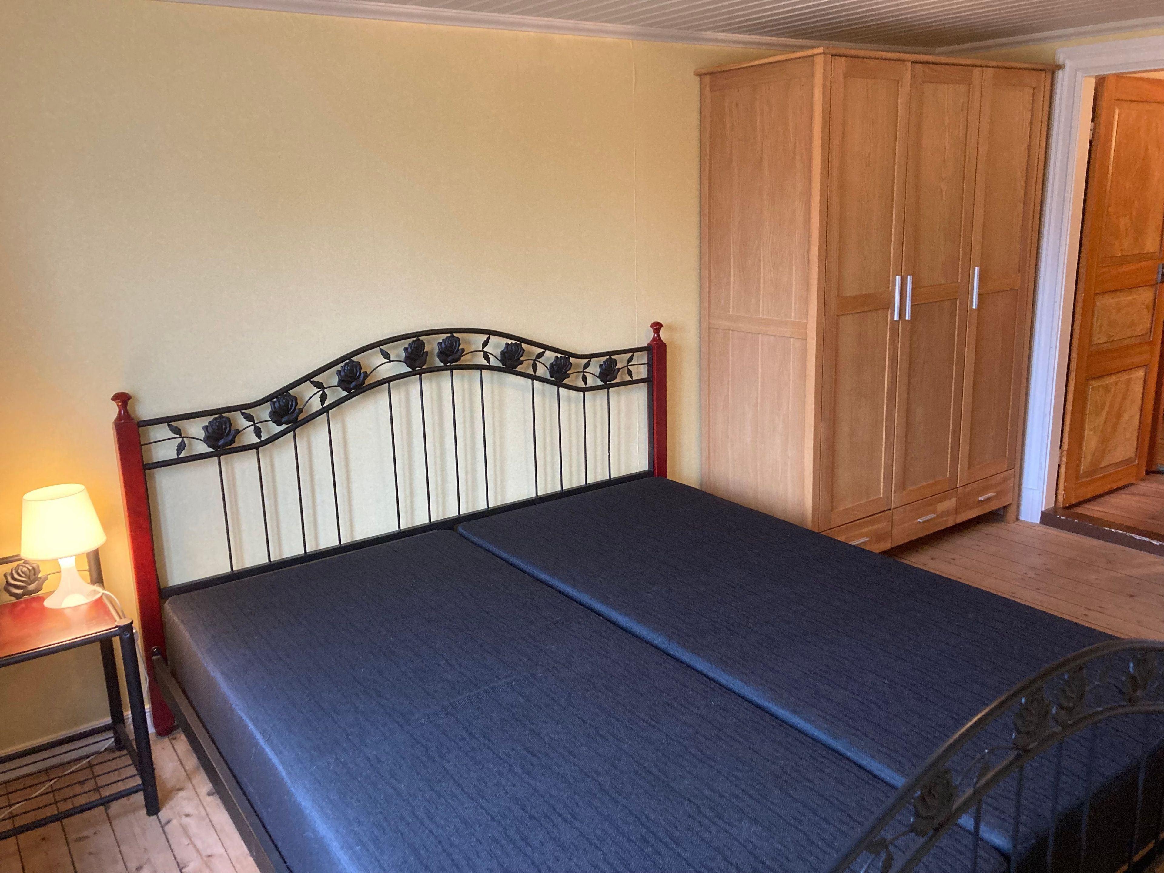 A single bed with a metal headboard, a wooden wardrobe, and a bedside table with a lamp.