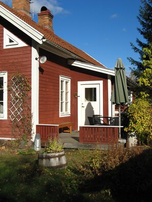 A red wooden house with a white door, a small porch with a bench, and an umbrella.