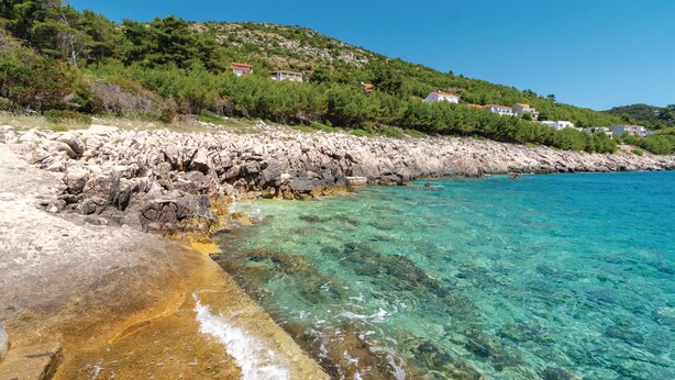 Un littoral rocheux bordé d’une eau turquoise et limpide, une plage de sable et des maisons construites sur une colline.