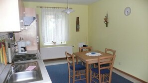 Uma cozinha compacta com pia, fogão e uma mesa de jantar com quatro cadeiras. Há uma janela com cortinas e um relógio na parede.
