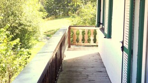 A wooden deck with a railing, a covered porch, and a view of greenery.