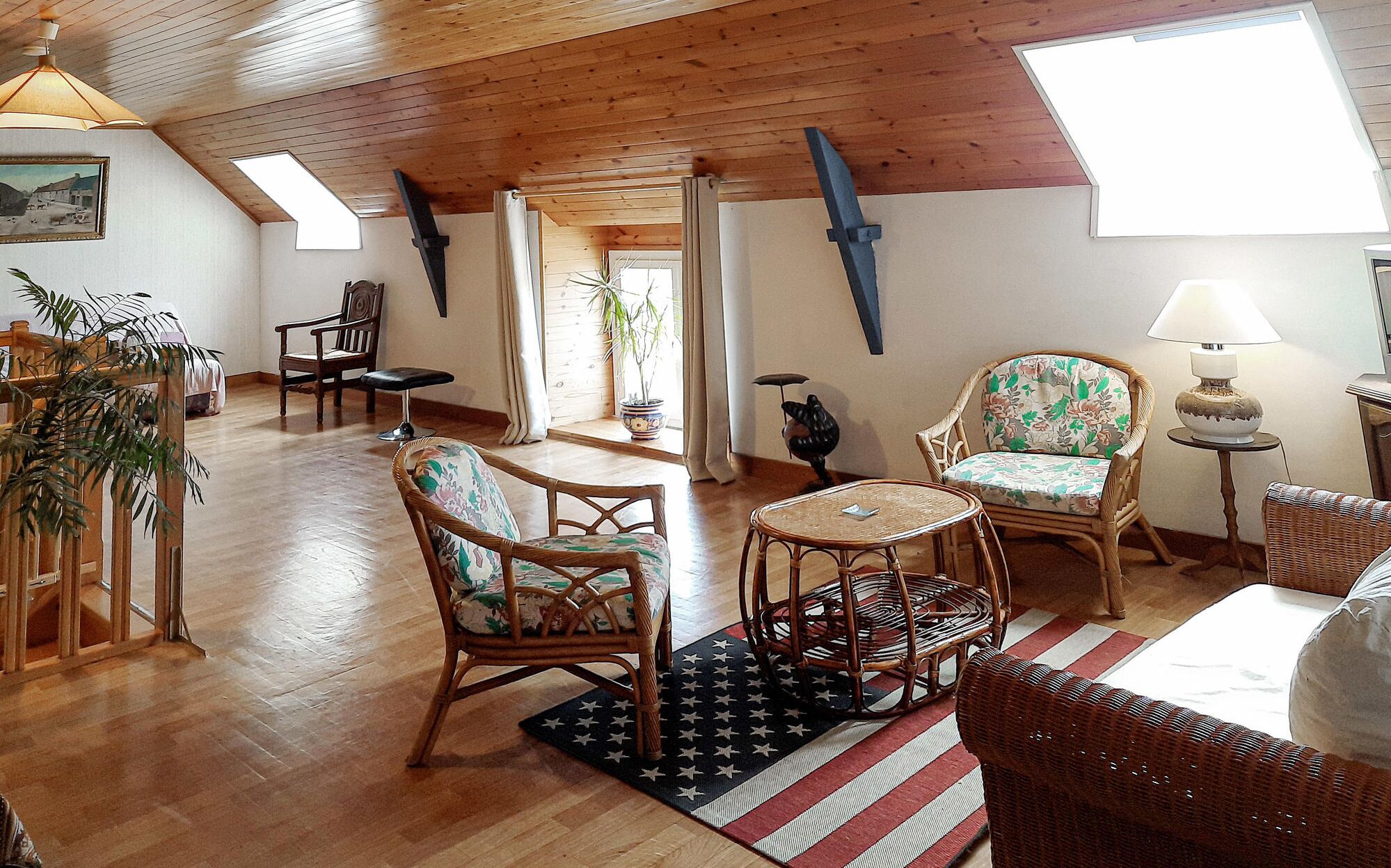 A cozy loft-style room with wooden floors, a high ceiling, and a skylight. There are wicker chairs, a small round table, and a lamp.