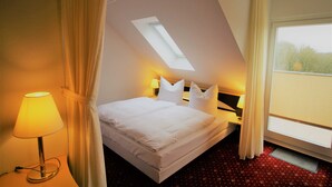 Une chambre d’hôtel comprenant un lit, une table de chevet, une lampe, une fenêtre avec des rideaux et une lucarne.