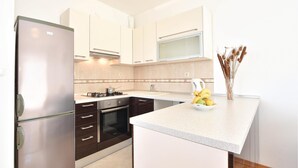A modern kitchen with white cabinets, a stainless steel refrigerator, a microwave, and a sink. There is a small island with a bowl of fruit and a vase of dried plants.