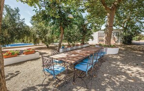 An outdoor dining area with a long wooden table and blue cushioned chairs, surrounded by trees and a pool. - Awesome home in Riudoms (Riudoms)