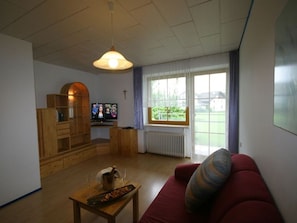 A living room with a wooden cabinet, a television, a red sofa, and a window with a view of greenery.