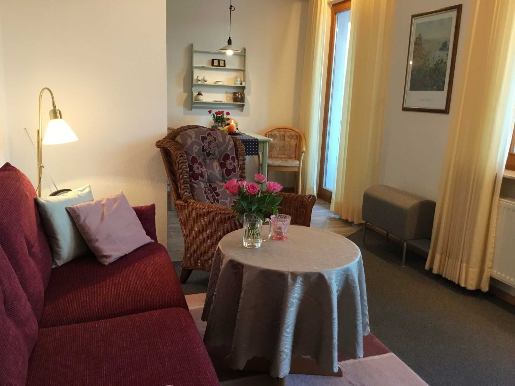 Ein gemütliches Wohnzimmer mit einem roten Sofa, einem kleinen runden Tisch mit Blumen in einer Vase, einem Korbsessel und einem Bücherregal.