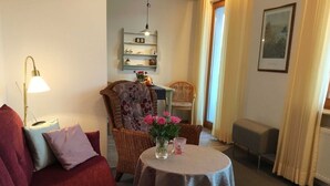 Un salon chaleureux avec un canapé rouge, une petite table ronde ornée d’un vase de fleurs, un fauteuil en osier et une bibliothèque.