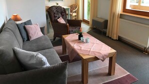 Un salon chaleureux avec un canapé gris, une table basse en bois et un fauteuil en osier. On y trouve une petite étagère avec des objets décoratifs et une fenêtre agrémentée de rideaux.