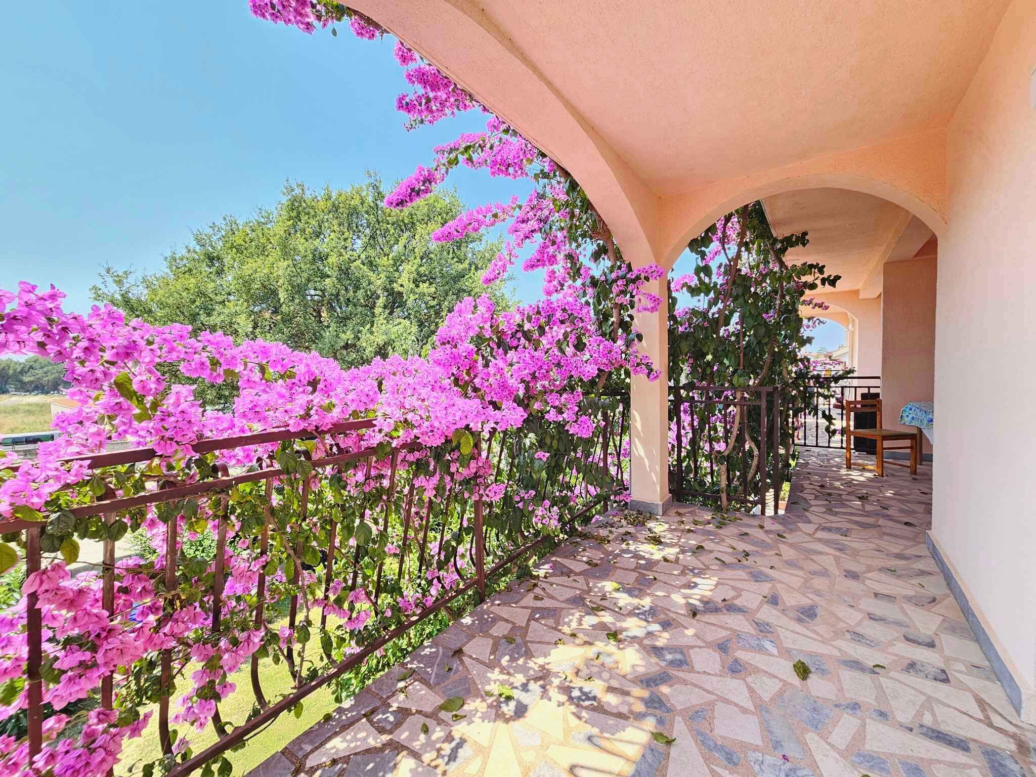 A balcony with a tiled floor and a metal railing, surrounded by pink flowers and greenery.