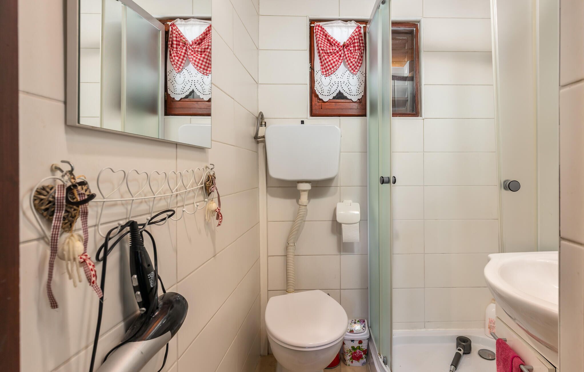 A small bathroom with a toilet, a shower, and a sink. There is a mirror above the toilet and a towel rack with towels and hairdryers.