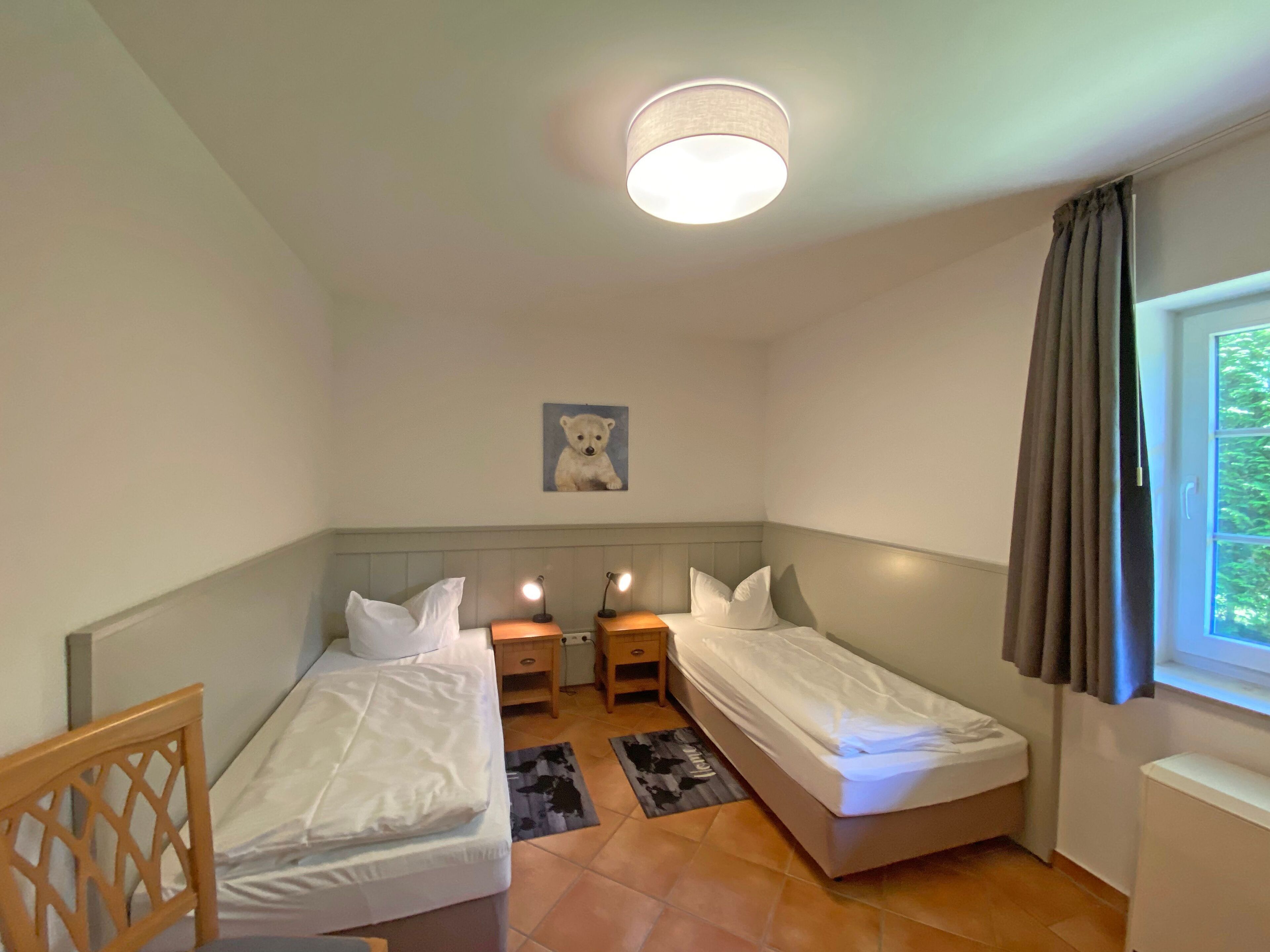A small, well-lit room with two single beds, a nightstand with lamps, a small rug, and a window with curtains.