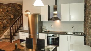 A modern kitchen with white cabinets, a stainless steel refrigerator, an oven, and a sink. There is a dining table with chairs and a pendant light above it.