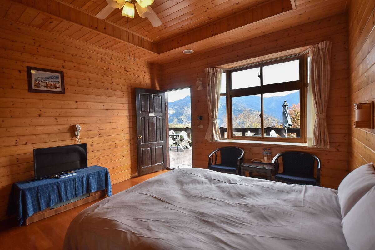 Habitación de cabaña de madera con una cama, un televisor sobre una mesa, un ventanal con vista a la montaña y una puerta que da al exterior.