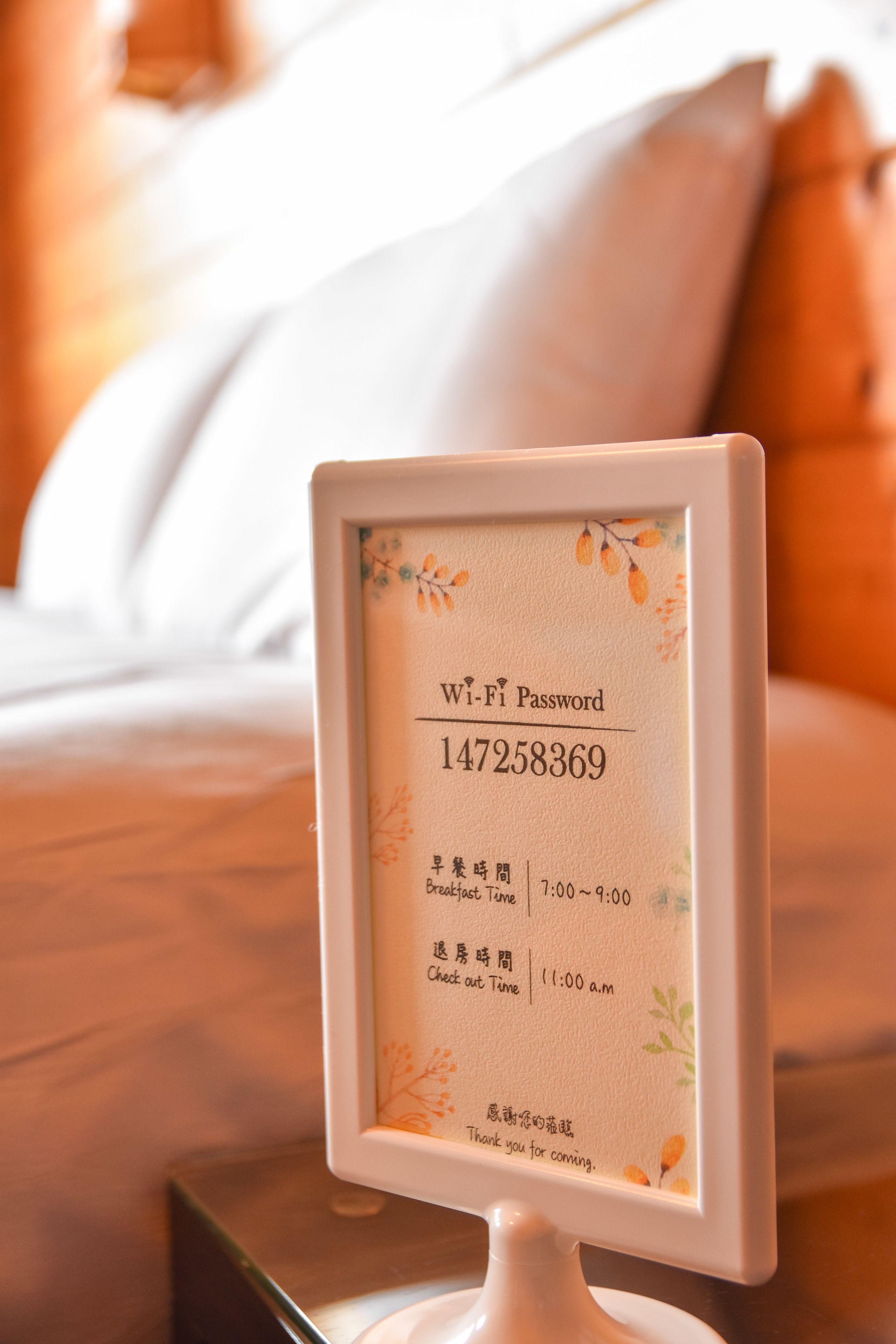 A framed Wi-Fi password sign with a floral border, placed on a bedside table in a hotel room.