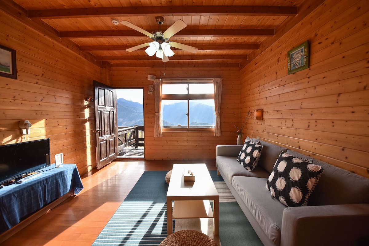 Interior de una cabaña de madera con sofá, televisión y vistas a las montañas a través de una ventana.