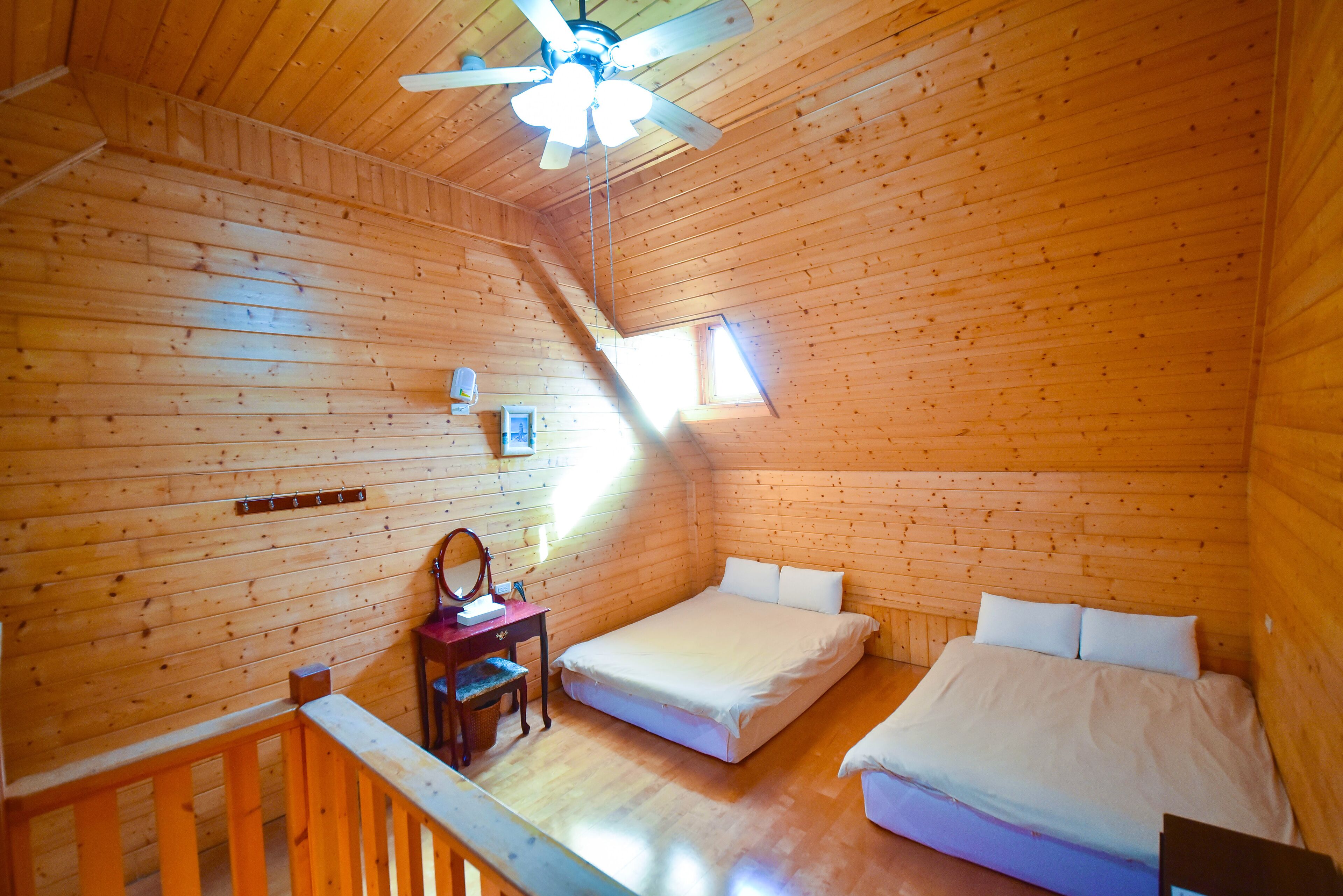 A wooden cabin bedroom with a sloped ceiling, a ceiling fan, a desk with a chair, and two beds.