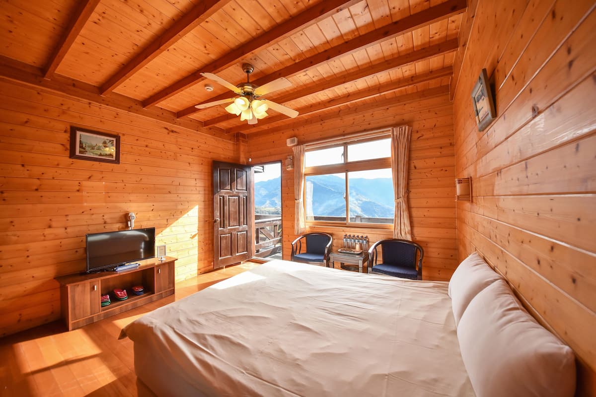 Un dormitorio de cabaña de madera con cama, televisor, ventilador de techo, ventana con vista a las montañas y puerta que da al exterior.