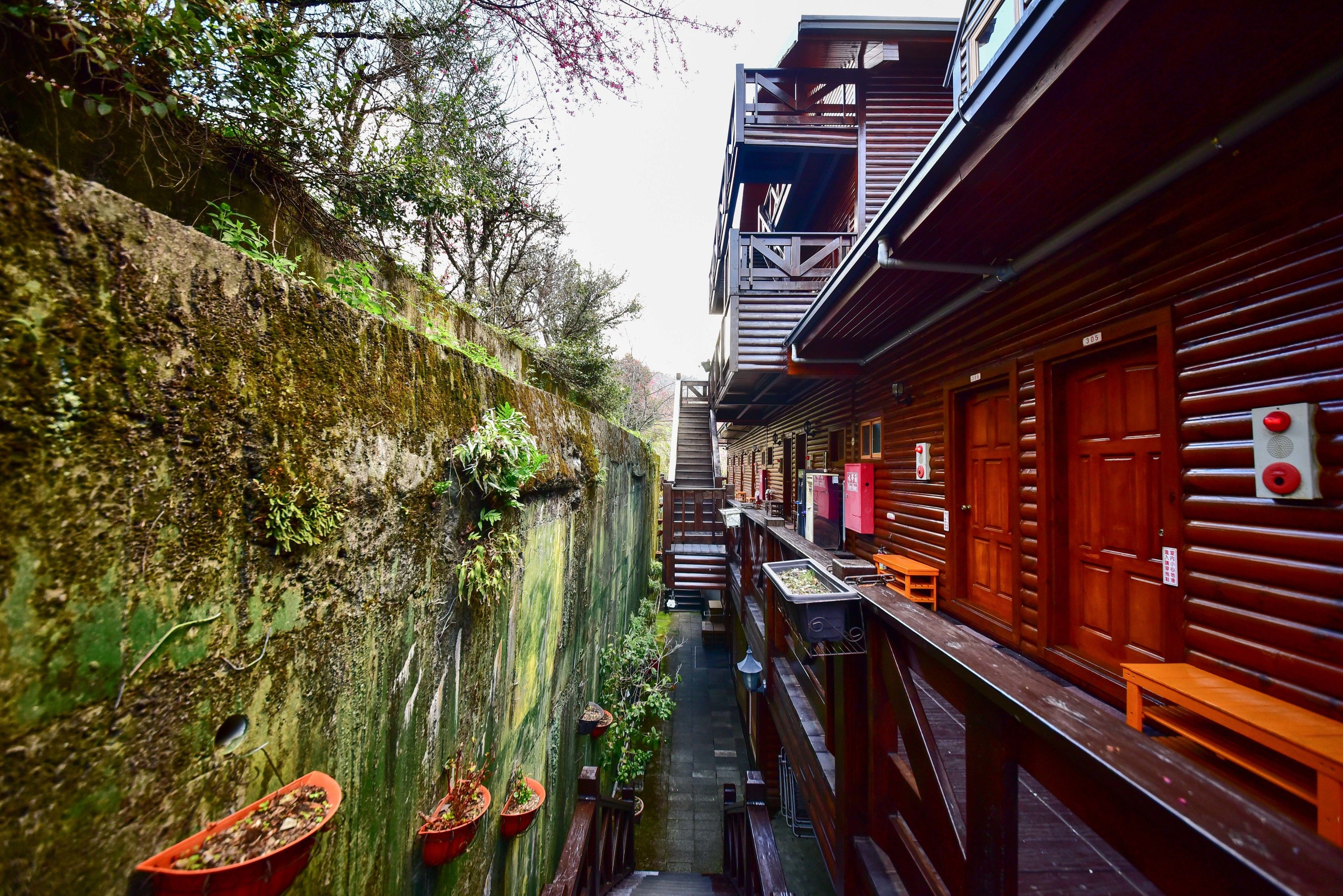 A narrow alleyway with wooden buildings on either side, potted plants, and a staircase leading to a second floor.