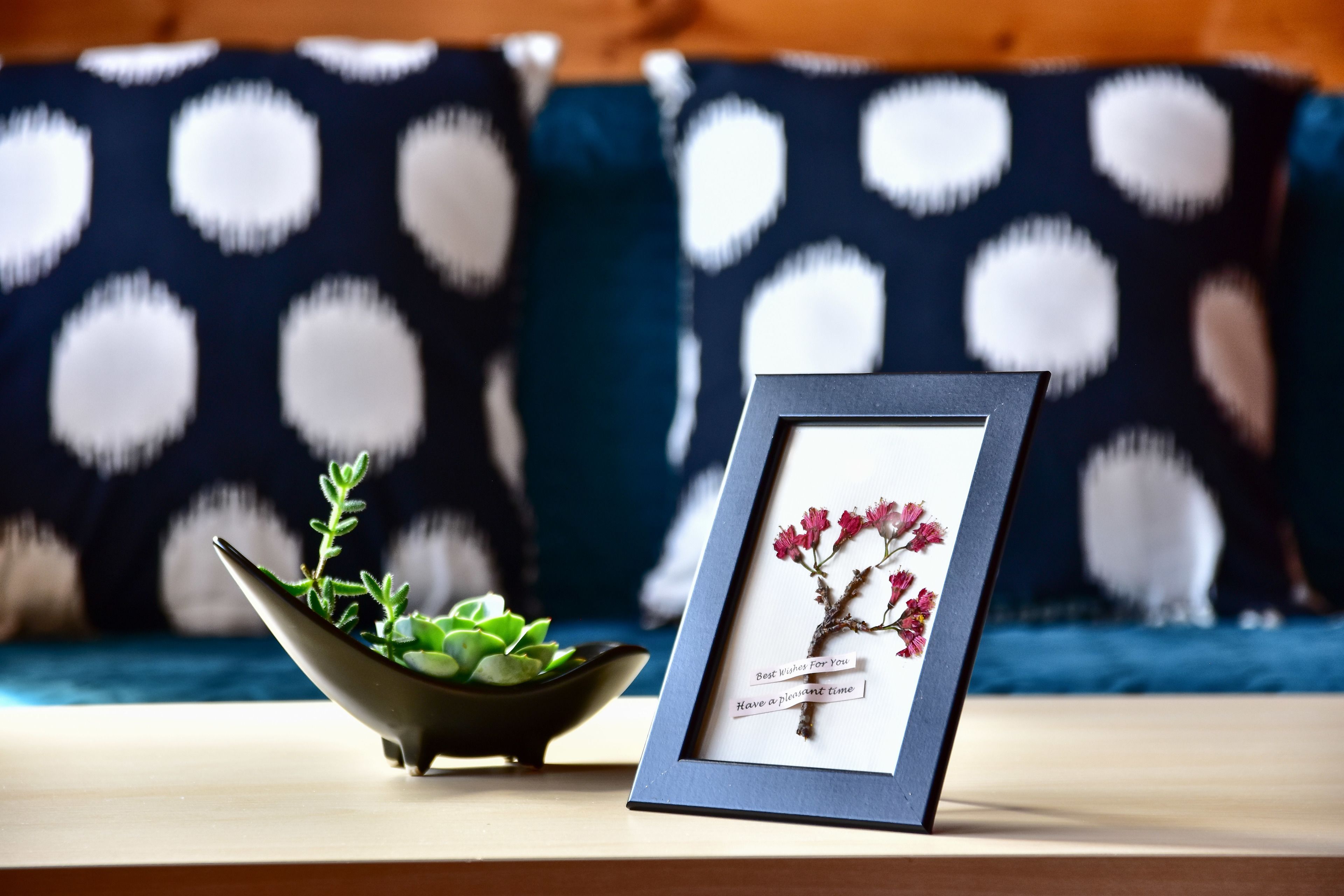 Un cuadro enmarco con flores sobre una mesa, junto a una pequeña planta en un cuenco, frente a un cojín estampado.