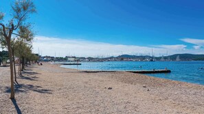 Uma praia de seixos com uma fileira de árvores e um porto no fundo.