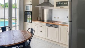 Une cuisine moderne équipée d’un four encastré, d’un micro-ondes, d’une hotte en acier inoxydable et d’un lave-vaisselle. On y trouve une table et des chaises en bois, et une porte vitrée donne sur une piscine.