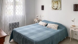 Une chambre à coucher avec un lit, deux tables de chevet, une lampe, une fenêtre avec des rideaux légers et un tableau représentant une rose accroché au mur.