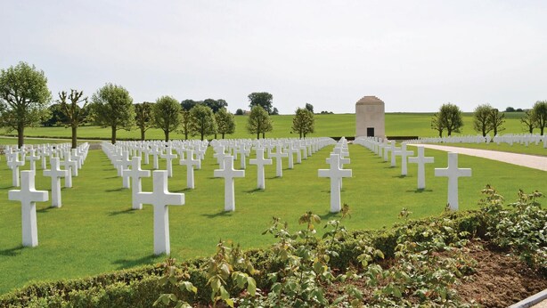 Un cimetière où de nombreuses croix blanches sont disposées en rangées, avec un monument central et une pelouse bien entretenue, agrémentée d’arbres.