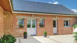 Un bâtiment en briques avec un toit à deux versants, deux portes d’entrée et des fenêtres avec des rideaux. Des plantes en pot sont disposées au niveau du sol.