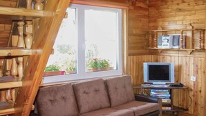 Interior acolhedor de uma cabana, com decoração em madeira, um sofá marrom, uma televisão sobre um suporte e uma janela com plantas em vasos.