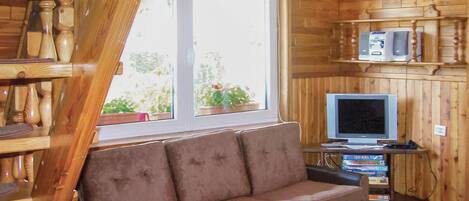 A cozy cabin interior with a wooden interior, a brown sofa, a television on a stand, and a window with potted plants.