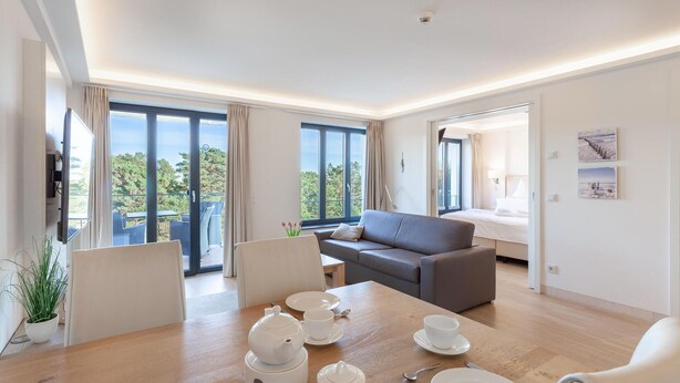 A modern living room with a sofa, dining table set with white tableware, and large windows offering a view of greenery. - 1 bedroom lovely apartment in Binz (Prora/Rügen)