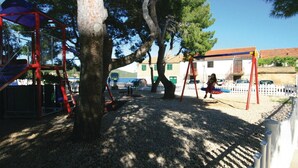 A playground with swings and climbing structures, surrounded by trees and a white fence.