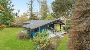 A small, blue cabin with a sloped roof, surrounded by a lush garden and dense trees. - 3 bedroom beautiful home in Idestrup (Idestrup)