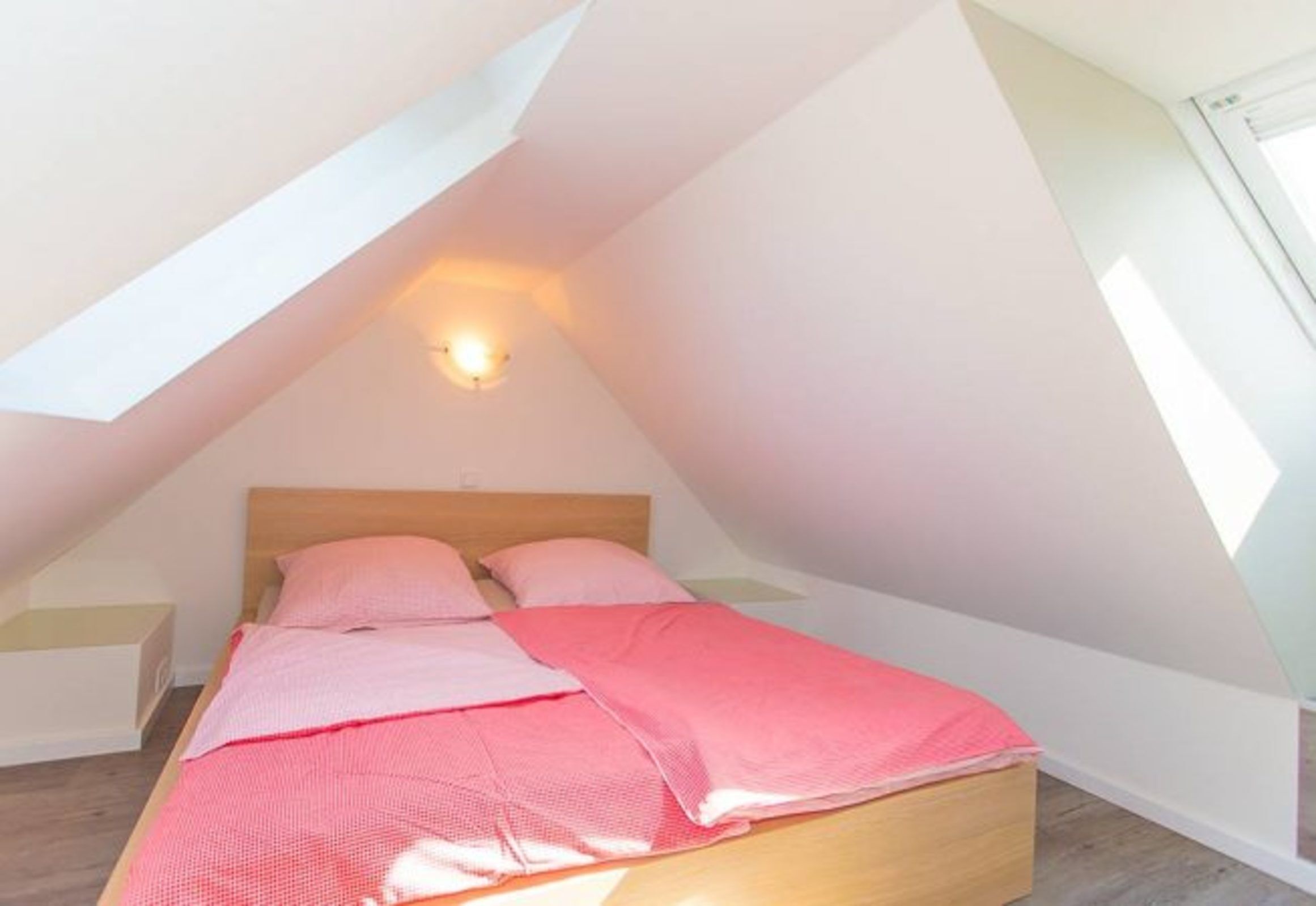 A bedroom with a sloped ceiling, a bed with pink bedding, and a wall-mounted light fixture.