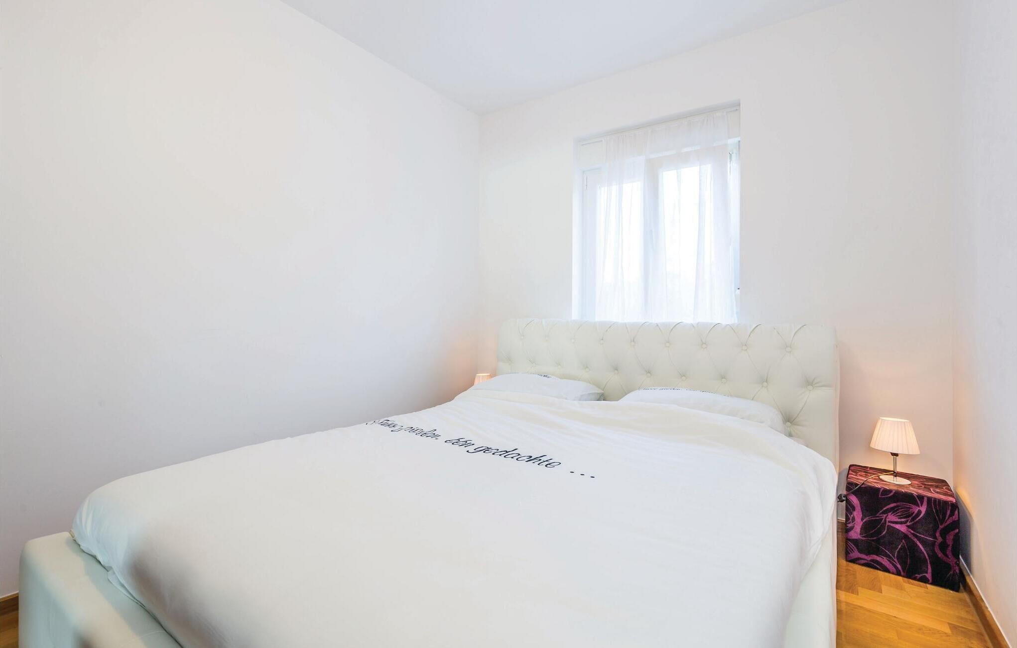 Quarto com cama, janela com cortinas leves, mesa de cabeceira com abajur e um tapete pequeno.