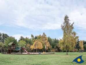 Un parque infantil con tobogán, columpios y estructura para escalar, en un parque con árboles y césped.