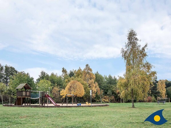 A playground with a slide, swing set, and climbing structure in a park with trees and a grassy field.
