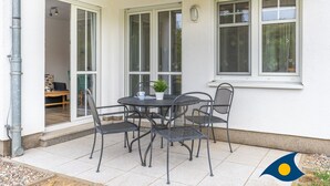 Un patio exterior con una mesa redonda y cuatro sillas, una planta en maceta y una ventana con vistas a árboles.