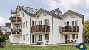 Un moderno edificio residencial de varios pisos con balcones, una zona de estar al aire libre cubierta y un césped bien cuidado.