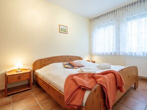 Une chambre à coucher avec un lit en bois, une table de chevet, une lampe, une fenêtre avec des rideaux légers et un tableau encadré accroché au mur.